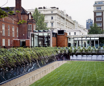 A grass covered patio bordered by a rectangular stone path. This lays surrounded by the red bricked Society building.