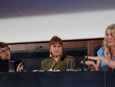 Three speakers on a conference panel sat behind a desk on a stage.
