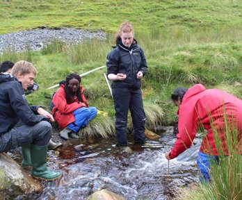 Three teachers sitting on the banks of a small, fast flowing stream and two standing in the water.