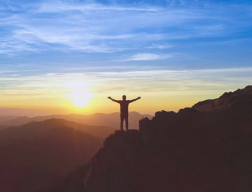 Person standing on a cliff with arms outstretched, in front of a sunset sky