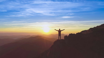 Person standing on a cliff with arms outstretched, in front of a sunset sky
