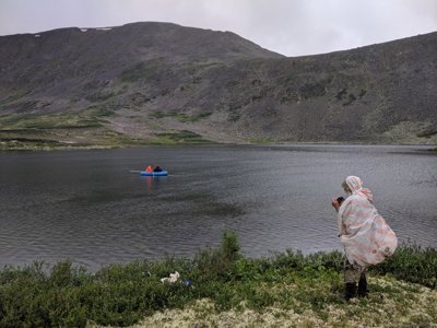 Two people sat in an inflatable boat on a lake with someone taking a photo from the bank of the lake. The lake is at the base of a steep-sided valley.