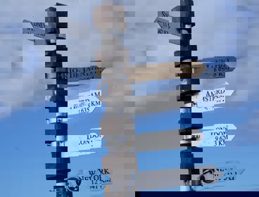 Brown and gray street sign pointing to various cities around the world with distance to them shown too