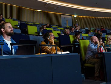 Audience members attentively listening in a modern lecture hall with tiered seating and green chairs.