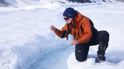 A field researcher bending down by a crack in a glacier holding a transparent tube and looking at the contents.