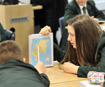 Children in a classroom. One holds a notebook with a world map, while another points at the map.