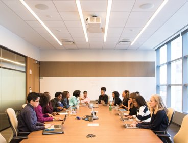 People sat around a long desk typing on laptops in a meeting
