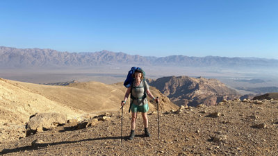 Person in trekking gear standing on top of a hill with a mountain range in the background.