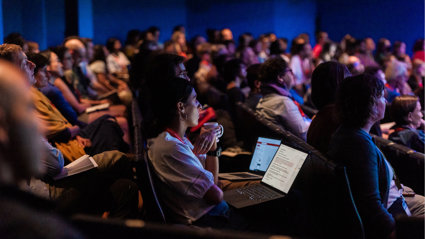 An audience listen to a presentation in a dark lecture hall.