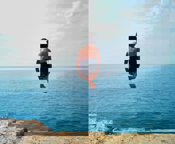 Person curled up, diving off a cliff into an blue ocean