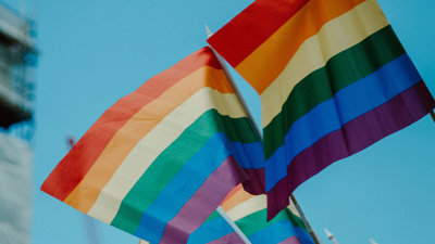 A series of LGBTQIA plus flags waving on front of a blurred backdrop which depicts a clear blue sky.