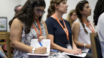 A person writing in a notebook while listening to a conference presentation, seated next to two other conference delegates