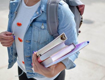 A student stands on a pavement holding two text books, a folder and notebook in one hand whilst holding on to their headphone cords with the other.