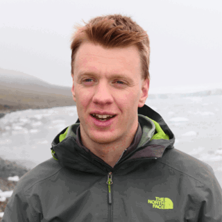 Headshot of Joshua Powell in the outdoors. In the background are a barren land and a sea with ice floating in it.