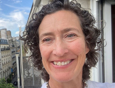 Person with curly hair wearing a white top standing on a balcony overlooking a cityscape with buildings and trees under a blue sky.
