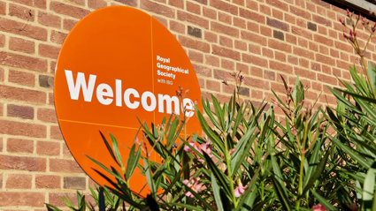 A circular orange sign reading 'Royal Geographical Society with IBG' and 'Welcome', next to a green plant with pink flowers.