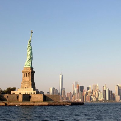 A view from the river of New York City, showing the Statue of Liberty and a skyscraper horizon. The sun is low in the sky so the light is soft
