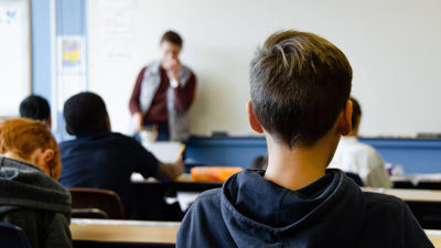 Several students faced towards a teacher at the front of a class room. The angle places one student in the foreground.