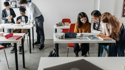 Six students congregate in two groups of three in a classroom with white walls and grey flooring. In both groups one student is seated at a desk whilst two others stand looking downward at papers on the desk. All the desks, whether empty or occupied, have paper, pencil cases and stationary sprawled over them.