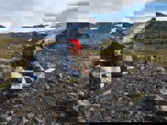 Researcher kneeling in a rocky landscape writing in a notebook. There is a glacier in the background.