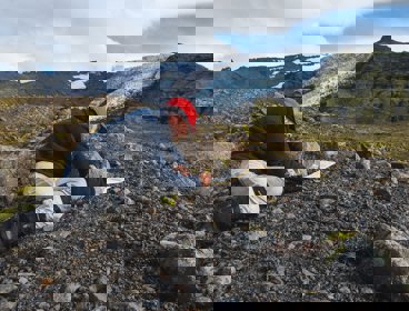 Researcher kneeling in a rocky landscape writing in a notebook. There is a glacier in the background.