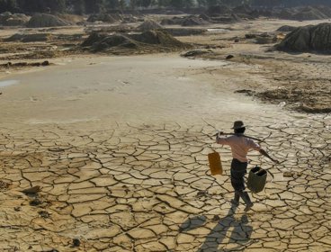 Person carrying water over dry lake with cracked earth.
