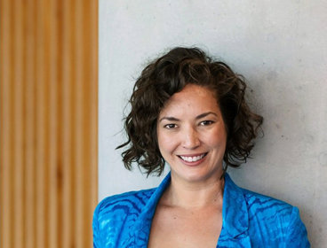 Person wearing a bright blue blazer standing with hands in pockets against a wall with wooden paneling.