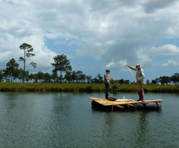 Two people stand on a wooden raft in the middle of a lake. The lake is surrounded by green grass land, sparsely dotted with trees.